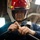 A person wearing a red firefighting helmet with yellow reflective strips and a face shield is working with a hose fitting or nozzle connector. They appear to be in an industrial or naval vessel interior environment, focused on connecting or adjusting a brass fitting on a dark hose. The setting suggests a damage control or firefighting drill/maintenance scenario.