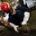 A naval personnel member wearing a red firefighting helmet with a face shield and white protective gloves is crouching down to handle a fire hose nozzle/strainer fitting on the deck of what appears to be a ship's hangar bay. Additional crew members in similar protective gear are visible in the background, along with aircraft and equipment. The scene appears to depict a damage control or firefighting drill aboard a naval vessel.