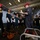 A military instructor wearing a red cap and coveralls with visible tattoos is briefing a group of personnel in firefighting gear inside what appears to be a naval vessel hangar bay. The trainees are equipped with red helmets, fire-resistant hoods, white gloves, and dark coveralls. Firefighting equipment including hoses and nozzles is visible on the deck floor.