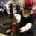 Military personnel, likely U.S. Navy sailors, are engaged in a firefighting or damage control training exercise aboard what appears to be a naval vessel. The foreground shows a sailor in full firefighting gear — red helmet, flash hood, and white gloves — operating a red fire hose valve or standpipe fitting. Several other sailors in navy coveralls with flash hoods and yellow boots stand observing in the background.