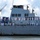 A large U.S. Navy warship, partially identified as 'USS LASSO' (or similar) on its hull, is docked at a port with numerous sailors in white dress uniforms manning the rails. A red and white ceremonial rope or ribbon is strung along the upper deck, suggesting a formal arrival, departure, or commissioning ceremony. The ship appears to be an Arleigh Burke-class guided-missile destroyer based on its superstructure design.