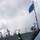 Naval personnel in white dress uniforms and blue hard hats are stationed along the bow of a gray military vessel. A large American flag (Union Jack/Navy Jack variant showing only the star field) is mounted on the bow flagstaff, decorated with a red and white striped pole. The scene appears to depict a formal naval ceremony, likely a commissioning, decommissioning, or port arrival event.