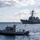 A U.S. Navy Harbor Patrol boat designated 'HP1' is operating in the foreground while a U.S. Navy Arleigh Burke-class guided-missile destroyer bearing hull number 82 is visible in the background. The destroyer appears to be either arriving or departing port, with sailors in dress whites manning the rails in a formal naval tradition. The scene takes place in a harbor under partly cloudy skies.
