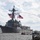 A U.S. Navy guided-missile destroyer is departing from a pier, flying multiple American flags and naval signal flags. Sailors in white uniforms are visible lining the deck in a traditional 'manning the rails' formation. A long red streamer extends from the ship toward the pier, likely a farewell or commissioning ribbon.