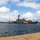 A U.S. Navy guided-missile destroyer is being assisted by two yellow-and-red harbor tugboats numbered 12 and 13 in a naval harbor. The warship is positioned between the two tugs, likely being maneuvered into or out of a berth. The scene is photographed from a pier with wooden bollards and mooring lines visible in the foreground.