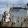Military personnel aboard a naval vessel are engaged in a mooring or docking operation, with one sailor in white dress uniform throwing a yellow heaving line with an orange monkey fist attached. Multiple sailors and personnel in camouflage uniforms and face masks are stationed along the ship's deck railing, which is fitted with safety netting. A port operations worker in an orange safety vest is visible on the dock below.