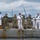 A group of U.S. Navy personnel are gathered on the deck of a naval vessel, all wearing face masks consistent with COVID-19 era protocols. The group includes officers in white dress uniforms and enlisted sailors in camouflage (NWU Type III) uniforms. One officer in white uniform appears to be speaking or giving remarks, possibly during a ceremony or briefing.