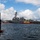 A large U.S. Navy gray warship, likely an Arleigh Burke-class guided-missile destroyer, is being assisted by two harbor tugboats numbered 12 and 13 painted in yellow and red. The scene is viewed from a wooden dock with mooring pilings in the foreground, suggesting the warship is either arriving at or departing from a naval base. Personnel in white uniforms are visible lining the deck of the destroyer, consistent with a formal arrival or departure ceremony.