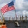 The image shows the USS Farragut, a U.S. Navy vessel, with personnel manning the rails in a formal naval ceremony. Sailors in both white dress uniforms and camouflage working uniforms are standing at attention along the ship's deck. A large American flag is prominently displayed and flying in the wind.