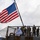 A large American flag flies prominently from a pole mounted on what appears to be a military naval vessel. Several uniformed personnel, wearing a mix of Navy working uniforms (camouflage) and dress whites, are visible on the deck behind a safety net railing. The personnel are wearing face masks, suggesting this was taken during the COVID-19 pandemic period.