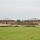 The image shows an active construction site featuring two nearly identical brick buildings with dark metal hip roofs, situated on a cleared and graded lot. Large mounds of excavated soil are piled between and around the structures, and erosion control silt fencing is visible along the perimeter. The site is bordered by a maintained grass field in the foreground and a tree line in the background.