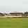 The image shows an active construction site between or adjacent to existing brick buildings with metal roofs, likely a school or institutional complex. The construction area features disturbed earth, a silt fence barrier, and a large dirt mound, indicating ongoing site preparation or expansion work. The foreground shows a well-maintained green lawn separated from the construction zone.