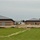 An active construction site featuring two nearly completed single-story brick buildings with dark metal hip roofs, situated on a graded sandy lot. The buildings appear to be institutional or commercial in nature, constructed with brown/tan concrete masonry units and horizontal accent bands. The foreground shows erosion control measures including silt fencing and landscape fabric, with a green grass field in the immediate foreground.