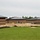 The image shows a commercial or institutional building complex under active construction, featuring dark metal standing-seam roofs and brown brick masonry walls. The surrounding grounds are largely bare earth with erosion control barriers (silt fencing) installed along the perimeter. The site appears to be in a late-stage exterior construction phase with landscaping not yet completed.