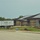 A commercial or institutional building is under active construction, featuring a brick exterior with horizontal banding and a dark metal standing-seam roof. An 'EB Insulation' delivery truck is parked on site, indicating insulation work is underway. The surrounding grounds show significant earthwork with mounds of displaced soil and an empty paved parking lot in the foreground.