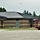 The image shows a single-story commercial or institutional building under construction or recently completed, featuring a dark metal standing-seam roof and brown/tan concrete block masonry walls. A red multi-level car hauler trailer is parked on the right side of the frame. The surrounding area includes a paved parking lot, freshly disturbed soil near the building foundation, and a backdrop of mature evergreen trees.