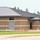 A commercial or institutional building is under active construction, featuring a concrete masonry unit (CMU) brick exterior with horizontal banding and a dark standing-seam metal roof. The structure appears nearly complete on the exterior but interior finishing work is still ongoing, as evidenced by unglazed window openings and disturbed soil around the perimeter. Construction safety equipment and at least one worker are visible on site.