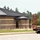 A commercial or institutional building is under active construction, featuring a brick exterior with a dark metal standing-seam roof. The structure appears nearly complete on the exterior but lacks finished landscaping and interior elements. A red flatbed/lowboy trailer is parked in the foreground right, likely associated with construction operations.