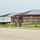 A commercial construction site showing a nearly completed brick building with a dark metal roof. An 'EB Insulation' delivery truck is parked on the left side of the building, indicating active interior work is still underway. The surrounding landscaping is partially installed with fresh grass, orange marker flags, and new signage posts.