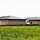 The image shows a large brick institutional or commercial building under construction, viewed from across an open grassy field. The structure features dark brick walls with light-colored horizontal banding, metal roofing, and minimal exterior doors visible. Construction debris and erosion control barriers (silt fencing) are present along the building's perimeter.