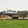 A construction site showing one or more brick buildings with metal hip roofs in an active phase of completion. Large piles of sand and gravel, heavy equipment, and stacked building materials are visible in front of the structures. The site is bordered by a grassy field in the foreground and a tree line in the background.