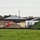 An active construction site surrounds what appears to be a newly built or recently renovated commercial or institutional building with dark metal roofing and brick masonry walls. The site shows significant earthwork in progress, with large soil mounds, construction materials, and temporary site infrastructure visible. A silt fence barrier runs along the perimeter, indicating erosion control measures are in place.