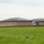 The image shows a large, single-story brick building under active construction, featuring a dark metal hip roof and tan/brown brick exterior walls. The building appears to be a commercial or institutional facility, possibly a school or community center, situated adjacent to an open grassy field. Construction activity is ongoing, with excavation work visible along the building's perimeter.