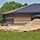 A construction site is visible in front of a newly built or nearly completed brick building with a dark metal standing-seam roof. Workers are present on the site, which contains piles of sand, gravel, construction materials, and equipment. The surrounding area includes green grass and mature trees, suggesting a suburban or semi-rural setting.