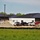 The image shows an active construction site adjacent to an existing building with a dark metal roof and brick/block construction. Multiple pickup trucks and work vehicles are parked on a dirt-covered area with large mounds of excavated soil or fill material. A silt fence barrier runs along the perimeter of the disturbed area, separating it from a green grassy field in the foreground.