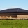 A large commercial or institutional building under construction, featuring a dark metal hip roof and brick masonry walls with decorative banding. The surrounding grounds are unfinished, with mounds of excavated soil and erosion control barriers (silt fencing) visible along the perimeter. A green grassy field occupies the foreground, and the sky is mostly clear and blue.
