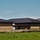 The image shows a newly constructed or nearly completed single-story commercial or institutional building with dark metal roofing and brick exterior walls featuring horizontal banding. Active construction or landscaping work is evident in the foreground, with disturbed soil and erosion control barriers (silt fencing) visible. The building is set against a clear blue sky with a green grass field in the foreground.