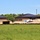 The image shows a commercial or institutional building complex under active construction, viewed from across a grassy field. The main structures appear largely complete with brick walls and dark metal hip roofs, while the surrounding grounds are still being developed with significant earthwork visible. Construction equipment, vehicles, and material stockpiles are present between the buildings.