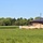 A rural construction site is visible at the edge of a large open green field, with a partially completed brick building featuring a dark metal roof on the right side of the frame. Construction materials including sand piles, lumber, and wrapped supplies are scattered around the work area. A treeline of mixed deciduous and coniferous trees forms the background under a clear blue sky.