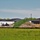 The image shows an active construction site adjacent to what appears to be a commercial or institutional building complex with dark metal roofing. Multiple mounds of soil and fill material are stockpiled along a silt fence barrier, separating the construction area from a green grassy field in the foreground. Several pickup trucks and a white trailer are visible within the construction zone.