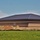 A large commercial or institutional building under construction, featuring a dark metal hip roof and multi-tone brick masonry walls. The surrounding area shows active earthwork with mounds of excavated soil and erosion control barriers (silt fencing) along the perimeter. The building appears structurally complete but the site work and landscaping are still in progress.