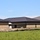 The image shows a newly constructed or recently completed single-story brick building with a dark metal standing-seam roof. The surrounding grounds appear to still be under development, with exposed soil mounds and black silt fencing/erosion control barriers visible along the perimeter. The building features a two-tone brick pattern with horizontal banding and is set against a green grassy field and clear blue sky.