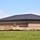 A large commercial or institutional building under construction, featuring a dark metal hip roof and brick masonry walls with decorative banding. The surrounding grounds are unfinished, with mounds of excavated soil and erosion control barriers visible in the foreground. A grassy field separates the viewer from the active construction site.