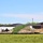 The image shows an active construction site adjacent to an existing building complex with dark metal roofing and brick exterior walls. Large mounds of excavated soil and sand are piled along a silt fence barrier, separating the construction zone from a green grassy field in the foreground. Several pickup trucks are visible near the buildings, indicating ongoing work activity.