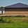 A large commercial or institutional building under active construction, featuring a dark metal standing-seam roof and concrete masonry unit (CMU) block walls with a decorative horizontal band. The construction site shows earthwork in progress, with disturbed soil, erosion control measures (black silt fence/plastic sheeting), and a delivery truck parked at the building. The surrounding area includes an open grassy field in the foreground and mature trees in the background.