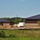 An active construction site featuring two partially completed brick buildings with dark metal roofs, surrounded by excavated earth mounds and construction equipment. The buildings appear to be commercial or institutional structures in a rural or semi-rural setting with wooded hills visible in the background. Construction is clearly ongoing, with exposed soil, silt fencing, and a contractor's trailer visible on site.