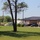 The image shows a suburban or rural commercial/institutional building with a dark metal roof and brick exterior, surrounded by a well-maintained green lawn. Several vehicles are parked in a lot adjacent to the building, and a white trailer with the text 'DEROUSSEAU' is visible, suggesting ongoing work or deliveries. The scene is photographed on a clear, sunny day with lush green trees in the background.