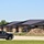 The image shows a newly constructed or nearly completed commercial or institutional building complex with dark metal roofing and brick masonry walls. Construction activity is evident from the large mounds of dirt/soil visible in front of the building. A gray RAM pickup truck is parked in the foreground left, and a red motorcycle is visible on the right side of the road.