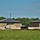 The image shows a commercial or institutional construction site featuring two nearly-completed brick buildings with dark metal standing-seam roofs. The foreground contains an unmowed grassy field with wildflowers, while the construction area between and around the buildings shows active site work with piled sand, equipment, and materials. The buildings appear structurally complete but surrounding site work and landscaping remain unfinished.