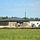 The image shows a commercial or institutional building under active construction, featuring a dark metal standing-seam roof and brick/block exterior walls. A green field occupies the foreground, separating the viewer from the construction site. Construction equipment, dirt piles, and parked vehicles are visible around the building perimeter.