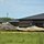 The image shows an active construction site adjacent to a large commercial or institutional building with dark brick walls and a black metal roof. Multiple piles of excavated soil and sand are visible in the foreground, along with construction equipment. The surrounding area includes green grass and wildflowers, suggesting a suburban or rural setting.