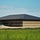 A large single-story commercial or institutional building under construction, featuring a two-tone brick exterior with dark brown upper courses and tan/buff lower courses, topped with a dark metal standing-seam hip roof. The surrounding grounds show active construction activity with exposed soil, sand piles, and erosion control barriers. A second similar structure is partially visible on the right edge of the frame.