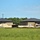 The image shows two nearly completed commercial or institutional buildings under active construction, viewed across an open grassy field. Both structures feature brick masonry walls with dark metal hip roofs and appear to be identical or mirror designs. Construction materials including sand/gravel piles, a yellow construction vehicle, and a small trailer are visible on the site.