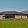 The image shows a newly constructed or recently completed single-story commercial or institutional building with brick exterior walls and dark metal roofing. Active landscaping and site work is underway, evidenced by exposed soil mounds and erosion control barriers (silt fencing) along the building perimeter. A second similar structure is visible in the background to the right.