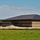 A large commercial or institutional building under active construction, featuring a dark metal hip roof and two-tone concrete block masonry walls. The surrounding grounds are unfinished with exposed soil, dirt piles, and erosion control barriers visible. Construction equipment and a contractor trailer are present on the left side of the site.