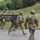Four soldiers in U.S. Army camouflage (OCP/MultiCam) uniforms are moving at a quick pace across a paved surface. Two of the soldiers are carrying what appears to be a military litter/stretcher between them, suggesting a casualty evacuation drill or training exercise. The setting appears to be a military installation or training facility with green hills and buildings visible in the background.